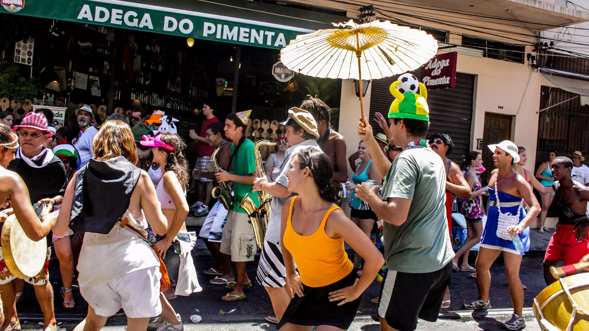 Pessoas fantasiadas tocando instrumentos e dançando em uma rua durante o Carnaval do Rio.